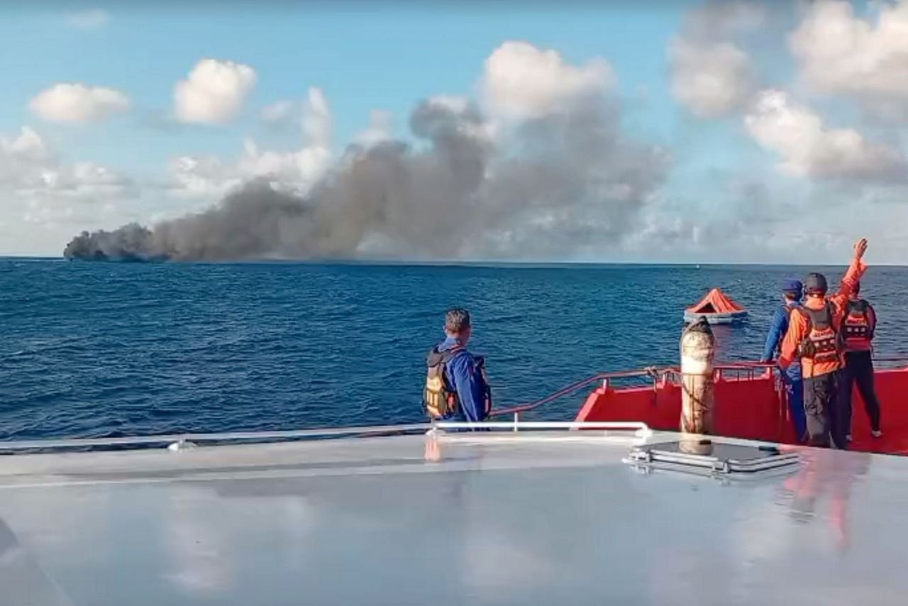 Rescuers in life jackets on a vessel watching smoke rise from a burning ferry, with a life raft in the water